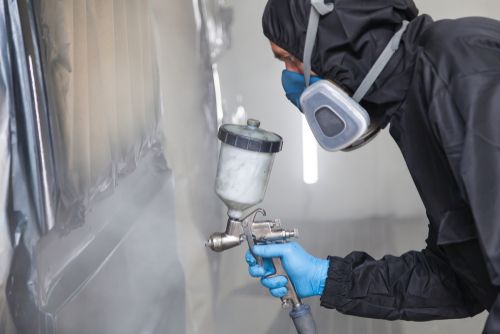 A male worker in jumpsuit and blue gloves paints with a spray gun a side part of the car body