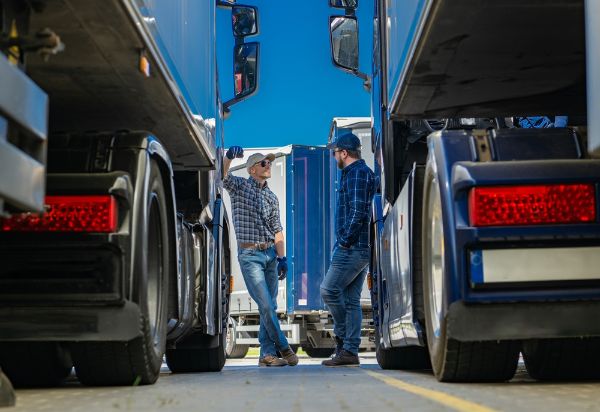 Two truckers are standing in front of a large semi truck. One man is holding a clipboard.