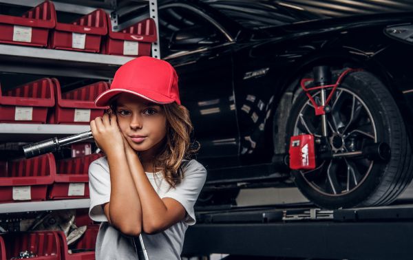 Shelf with tools, shiny car and little girl in hat with big wrench in hands.
