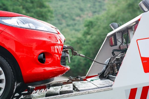 Close-up of a red car being towed by a tow truck on a rainy day with a green forest background.