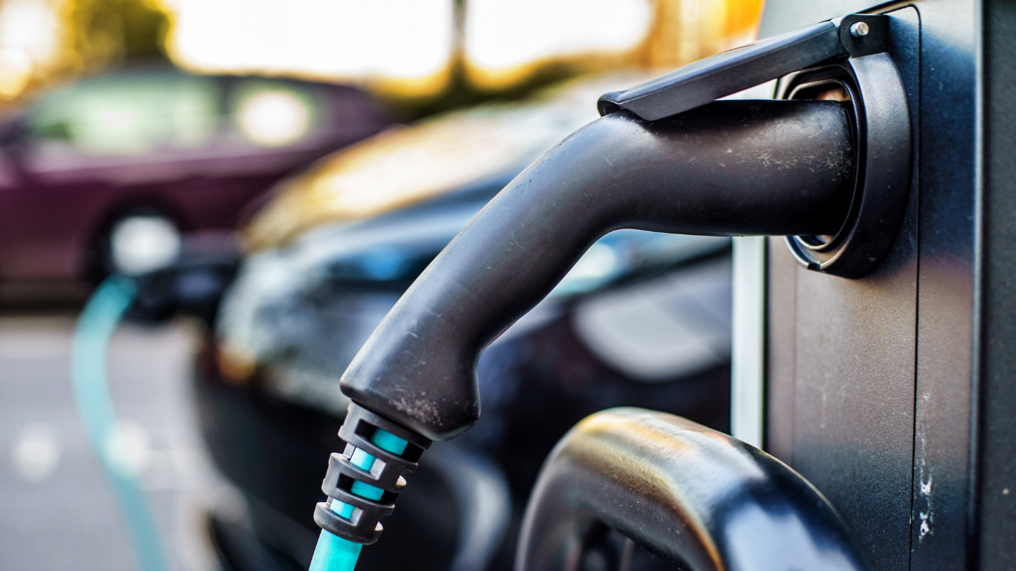 A close-up image of an EV charger plug with other EVs behind it.