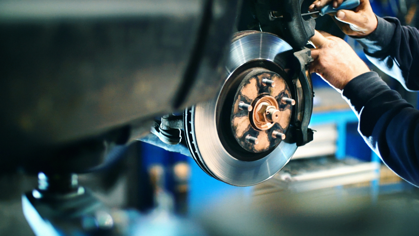 A close-up of a car brake being worked on by a mechanic who holds a blue-handled tool.