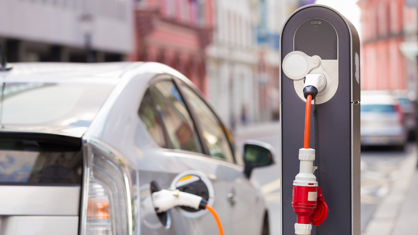 A white hybrid car is parked on a downtown street, it is plugged into a recharging station.