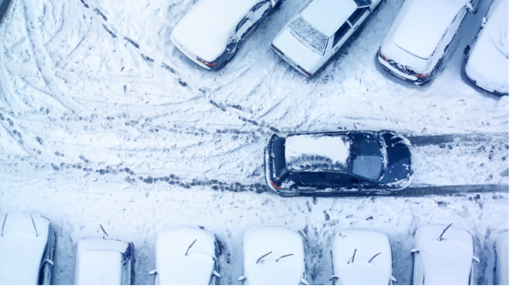 A wintery parking lot covered in snow with a car driving.